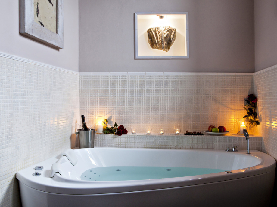 White corner soaking tub with candles, roses, and a champagne bucket on a tiled ledge in a spa-like bathroom.