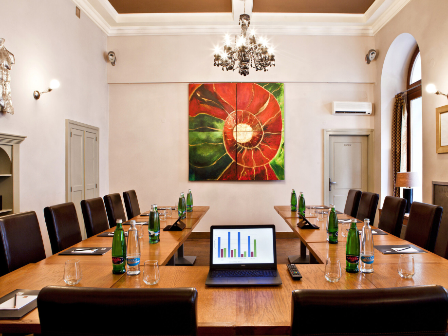 Conference room with a long wooden table, laptop showing bar charts, and water bottles in front of brown chairs under a chandelier.