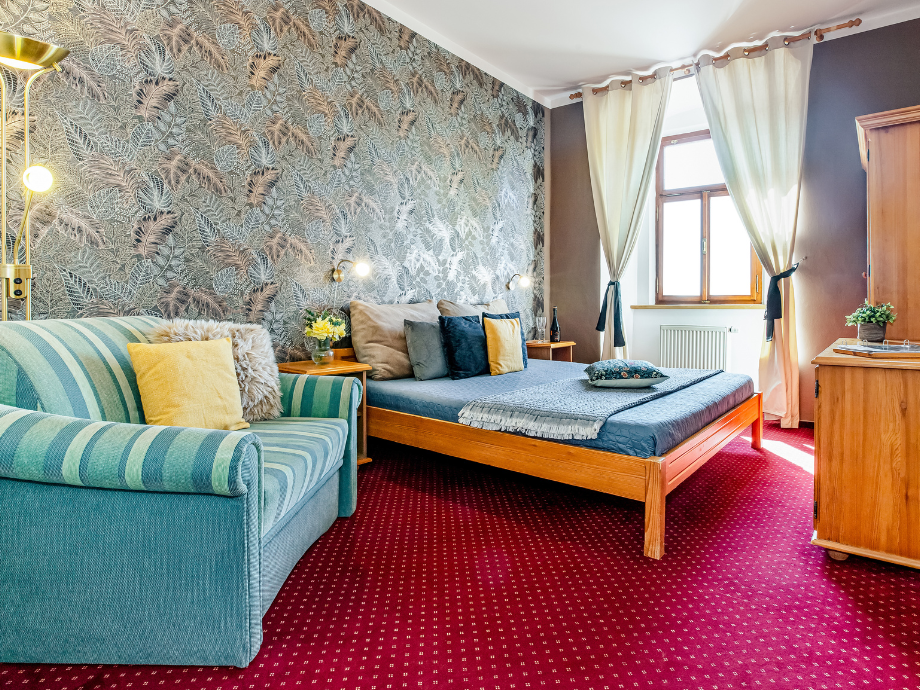 Hotel room with a wooden-framed bed, blue bedding, and a green striped sofa against a botanical wallpapered wall. A window with cream curtains and warm lighting complete the scene.