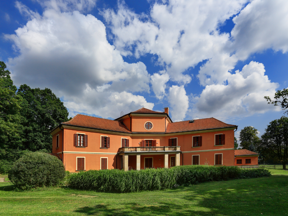 Orange two-story villa with a red-tiled roof and central balcony set in a well-kept lawn under a blue sky with white clouds, surrounded by trees and hedges.