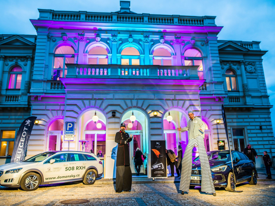 Historic building facade lit in blue and magenta, arched windows and balconies, with a white branded car and a striped-stilt performer outside as people enter.