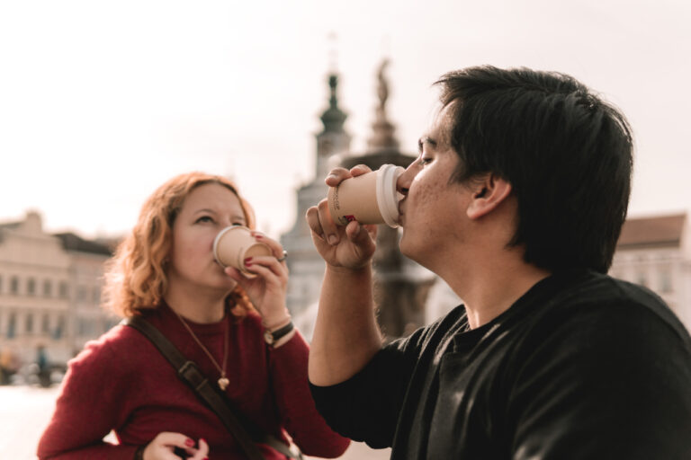 Two friends in an urban square enjoy coffee from disposable cups, facing each other as they sip outdoors.