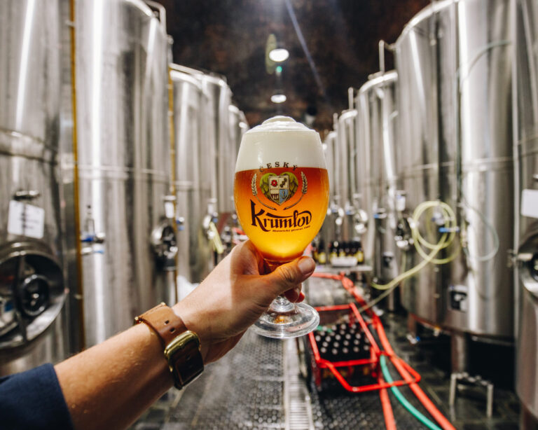 Glass of amber beer held up in a brewery with tall stainless steel fermentation tanks in the background; night setting.
