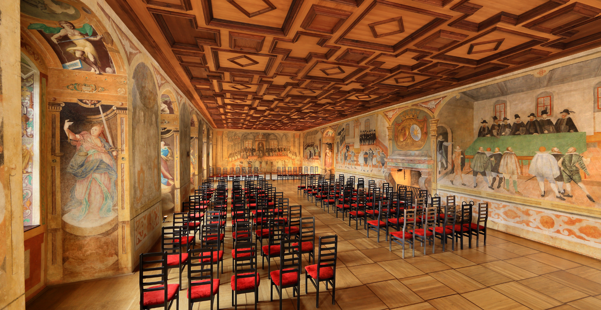 Historic frescoed lecture hall with a carved wooden coffered ceiling and long muraled walls, lined with rows of black chairs with red seats.