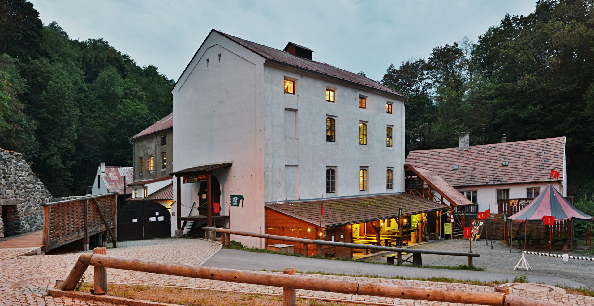 Three-story white building with lit windows in a cobblestoned courtyard, surrounded by trees and a wooden fence.