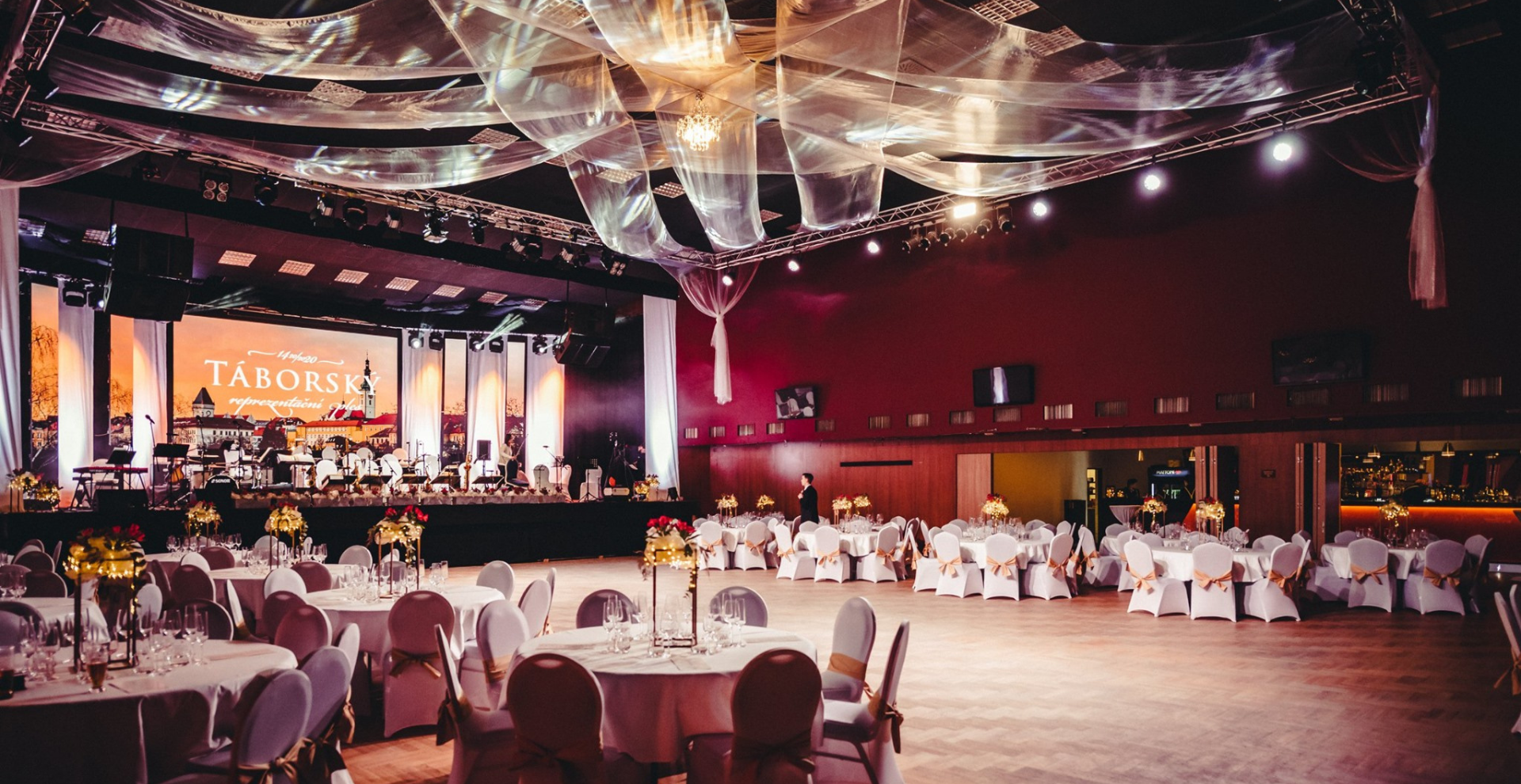 Elegant banquet hall with a live band on stage, velvet lighting, and draped ceiling decorations over round tables set with candles and gold bows.
