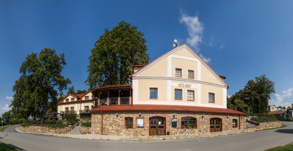 Hotel Gold: pale yellow building with white trim and red roof, stone base along a curved street with tall trees against a blue sky and a clear day.