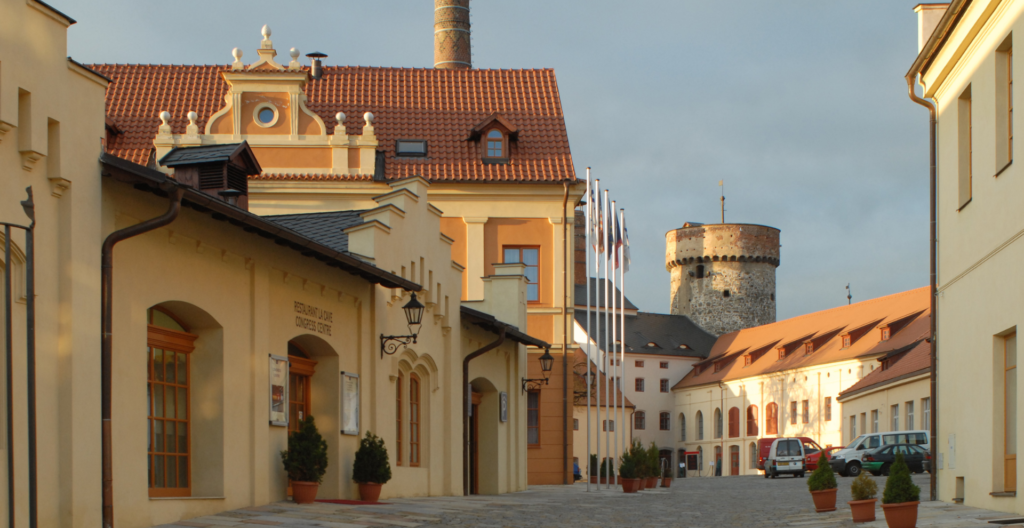 Sunlit European street lined with pastel buildings, red-tiled roofs, flagpoles, and a historic round stone tower in the background; parked cars along the cobblestone street.