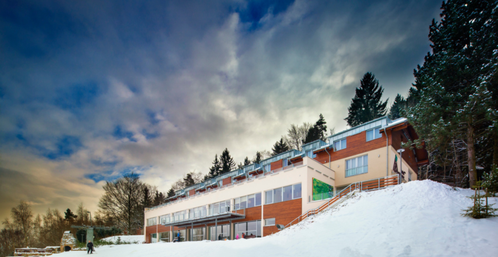 Modern, multi-story building on a snowy hillside with evergreen trees and a dramatic blue-gray sky.