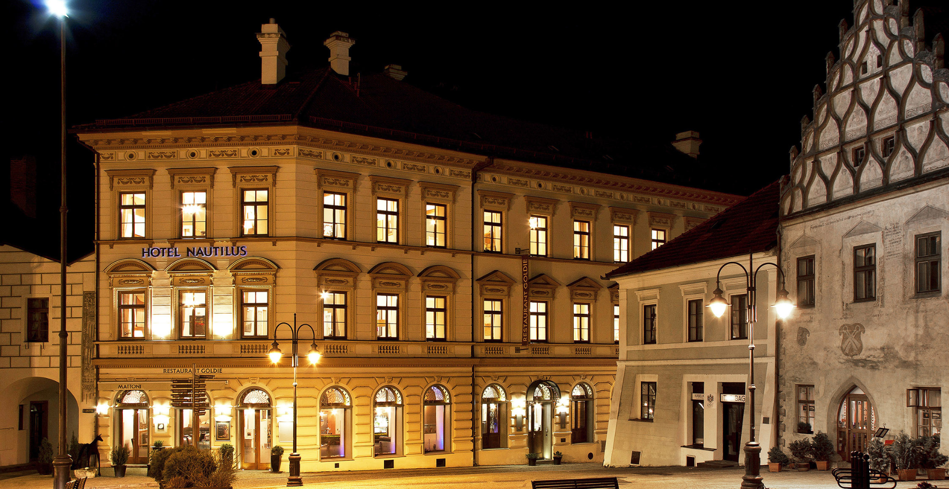 Night view of a lit historic building: Hotel Nautilius with warm interior lights and street lamps along a cobblestone square