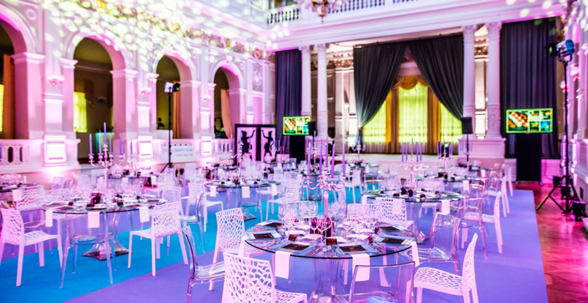 Banquet hall set for a gala: round tables with glassware and white chairs, pink lighting, and a stage with dark curtains.